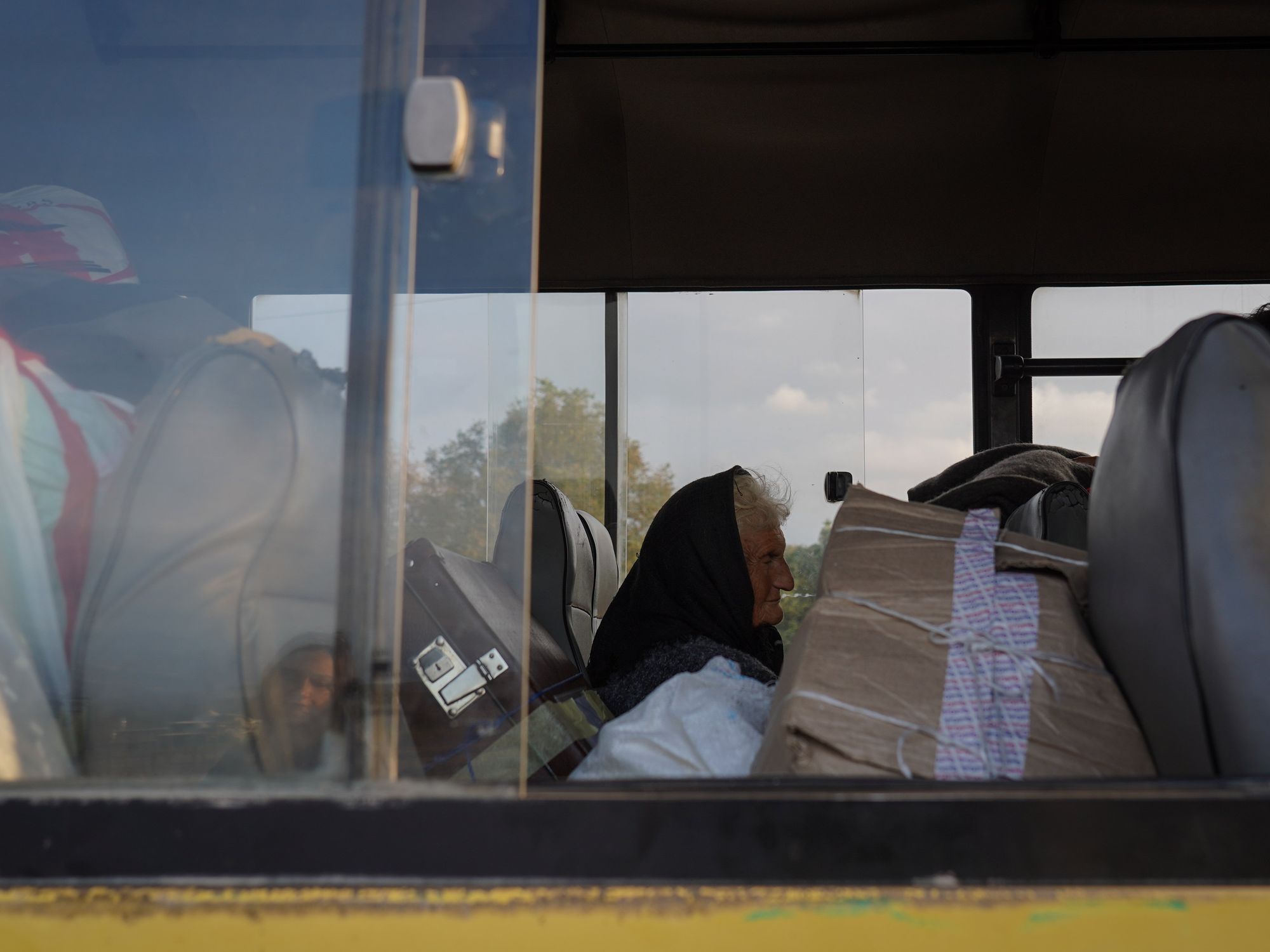 An elderly Women is seen seated in a bus surrounded by luggage overflowing inside the vehicle.