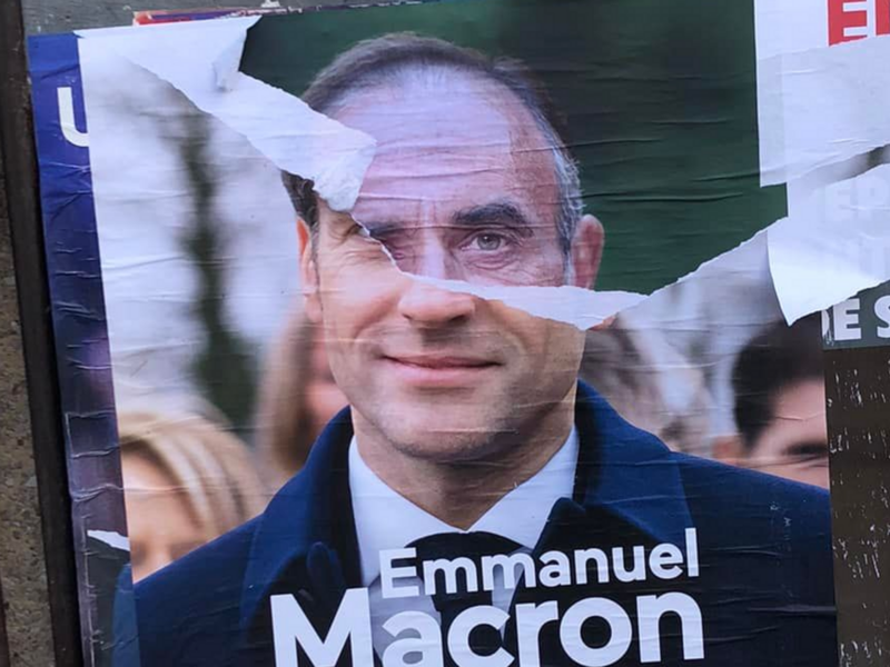 An electoral poster showing Eric Zemmour's head showing from beneath a torn campaign poster of Emmanuel Macron.