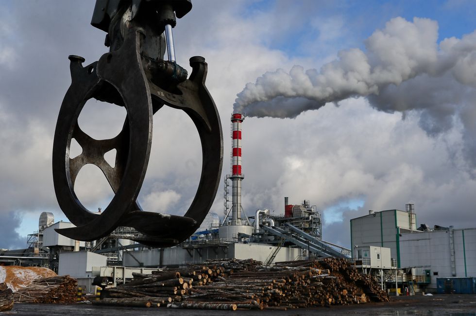 An industrial wood crane is seen at the front of the picture, with a large stack of wood piled in front of a factory with a smokestack in the distance.