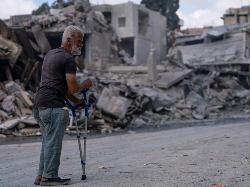 An old man walks slowly through the ruins of destroyed buildings, reflecting the weight of loss and vulnerability.