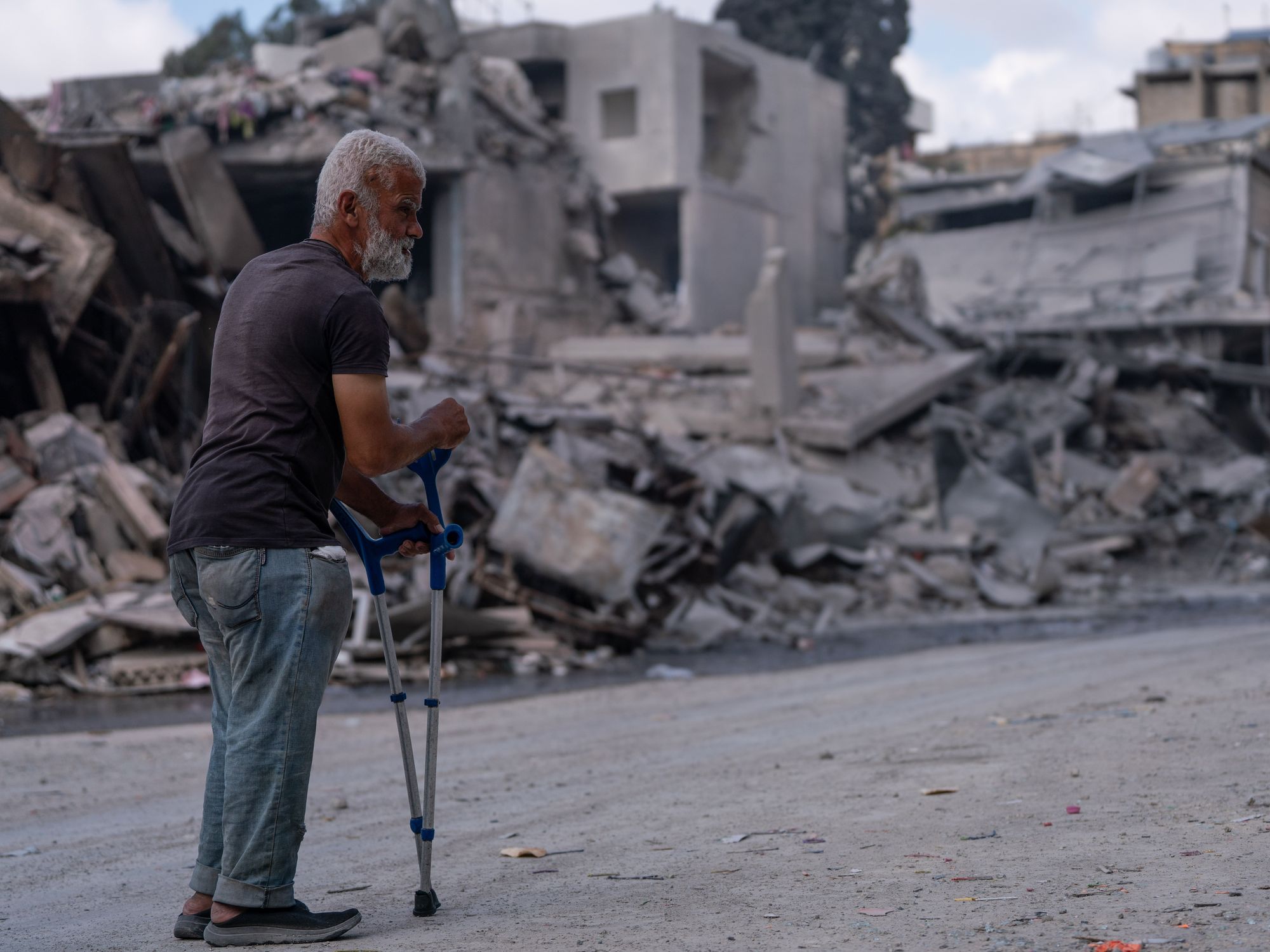 An old man walks slowly through the ruins of destroyed buildings, reflecting the weight of loss and vulnerability.