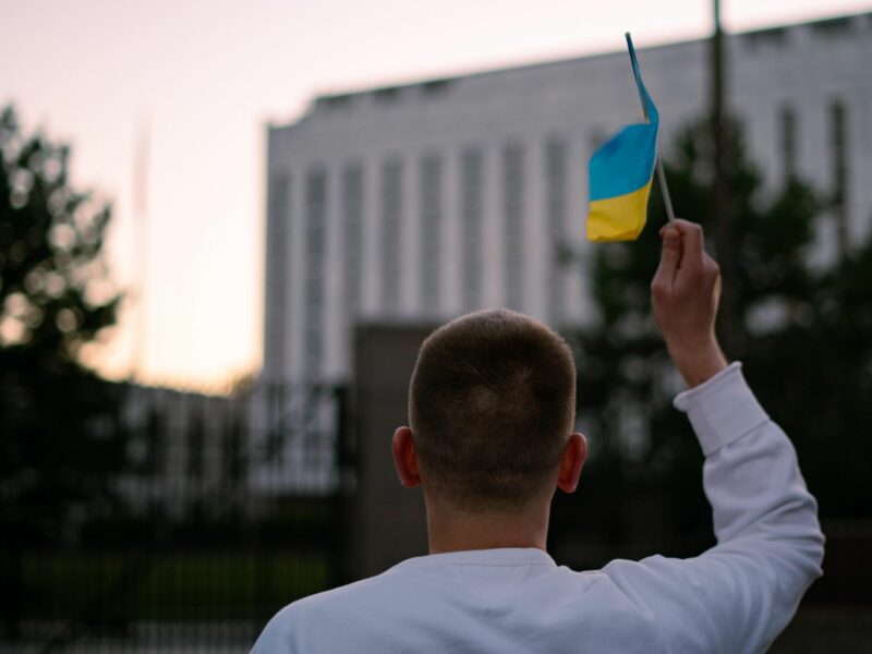 back vew of a man holding up and waving a little ukrainian flag in front of the Russian Embassy in Washington
