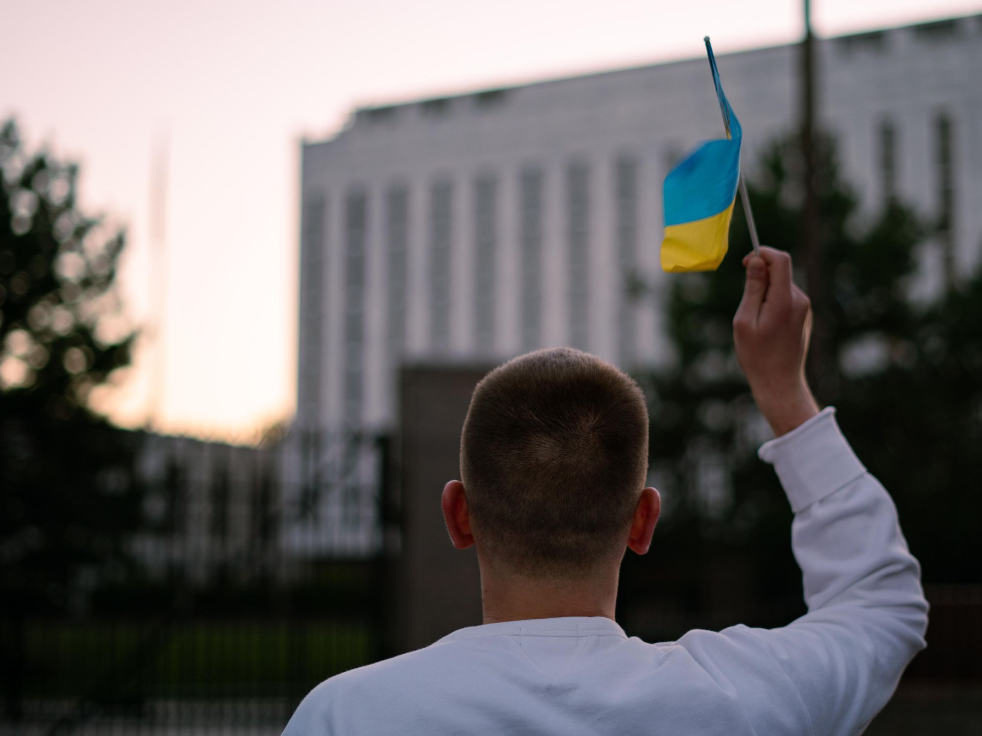 back vew of a man holding up and waving a little ukrainian flag in front of the Russian Embassy in Washington