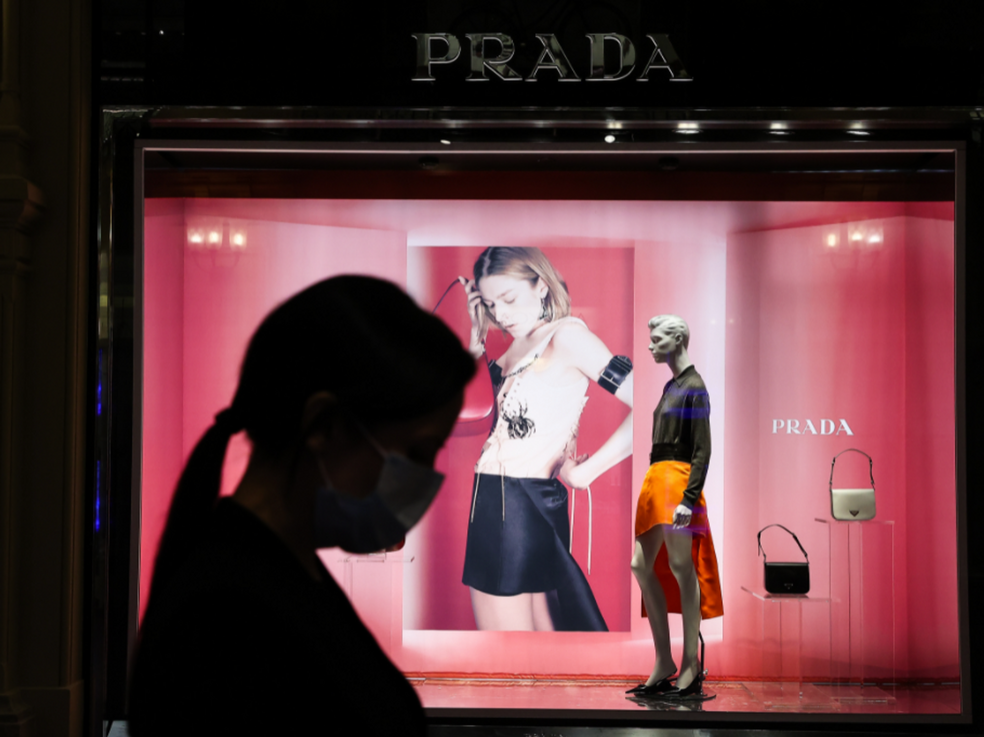 Backlit photo of a woman wearing a COVID mask passing by a PRADA shop window at the GUM luxury department store in Moscow​