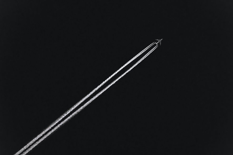 Black-and-white photo of an ariplane shot from below flying across the sky and leaving condensation trails