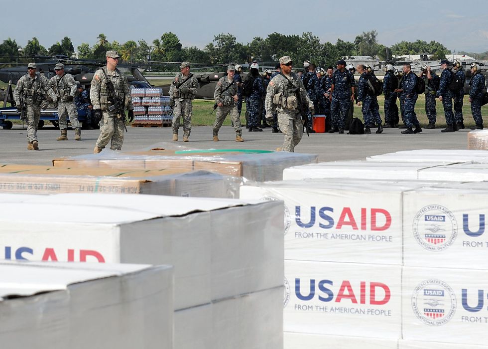 Boxes labeled USAID sit in the foreground while American military members file in the background.