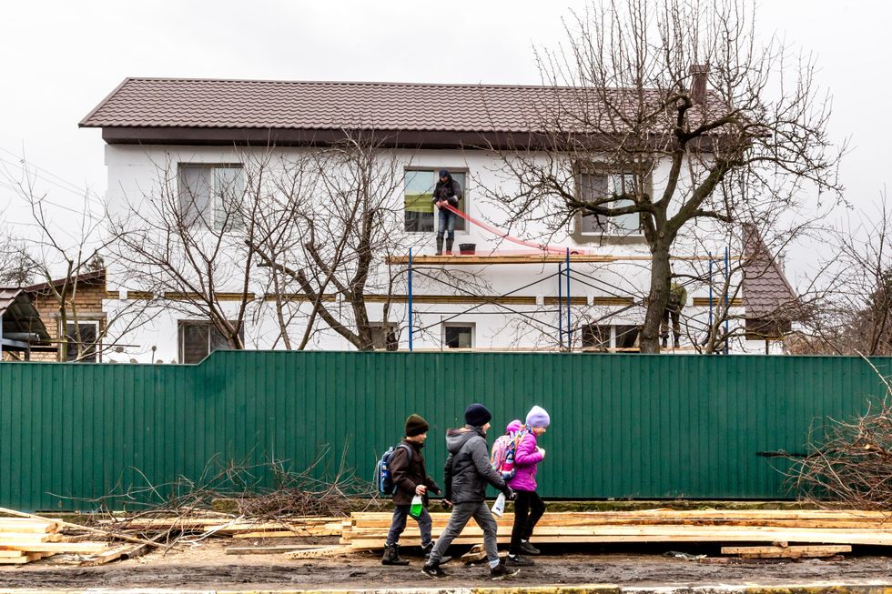 Children walk past woods on the pavement as builders renovate the roof of a bombarded house in Bucha.