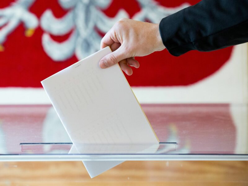 Close up on a hand putting a vote in a ballot box, with the Polish flag as backdrop