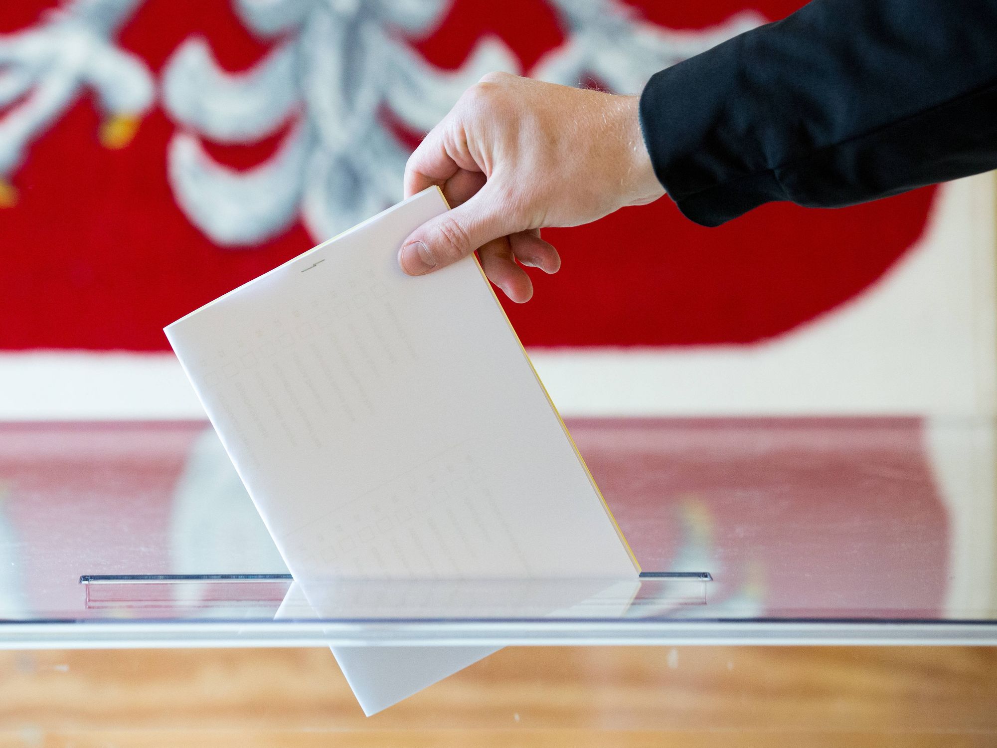 Close up on a hand putting a vote in a ballot box, with the Polish flag as backdrop