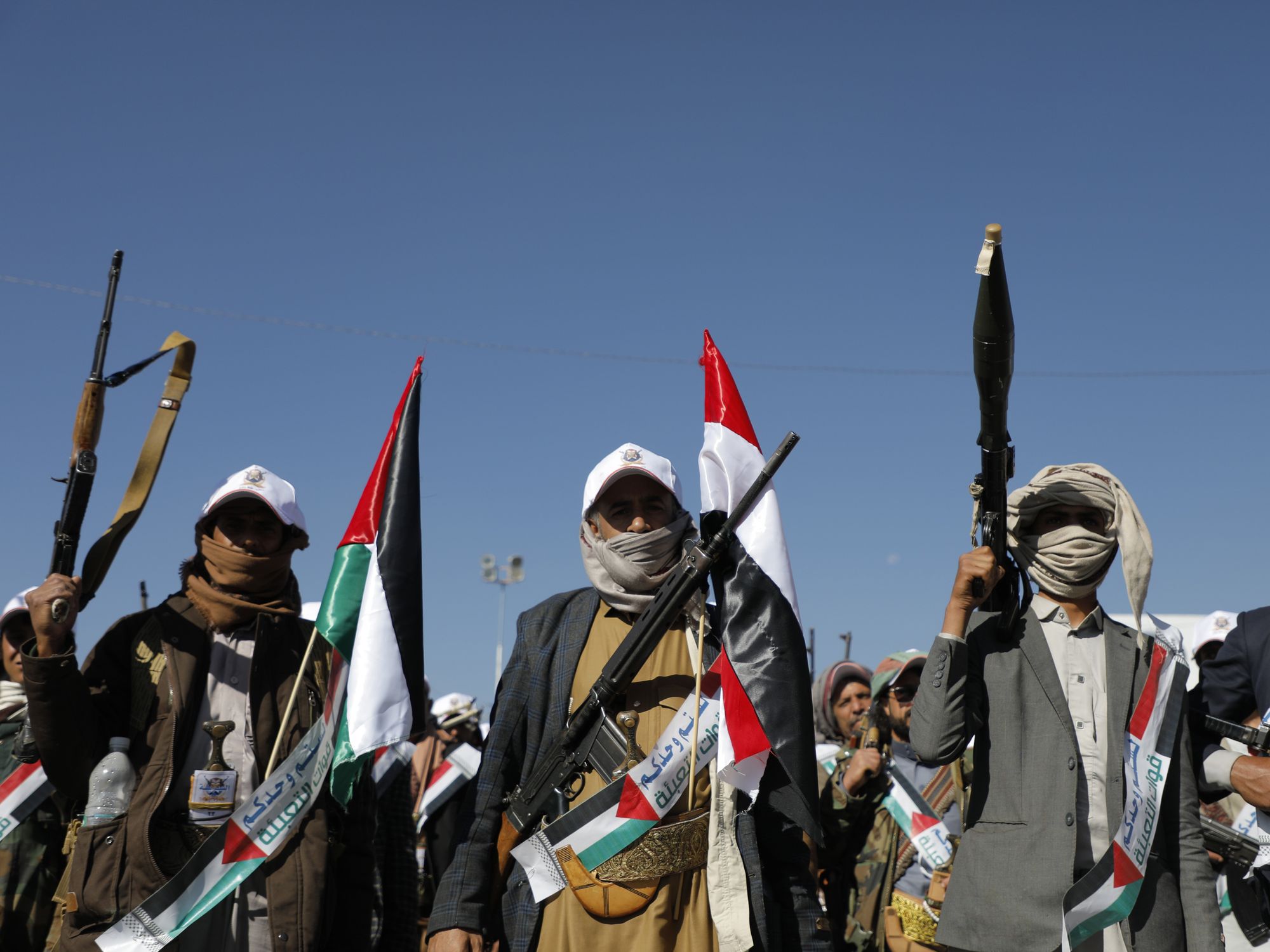 Crowd of newly recruied Houthi rebels participating in a military parade carrying weapons and rocket launchers. Holding and wearing both the Palestine flag and the Yemen flag.