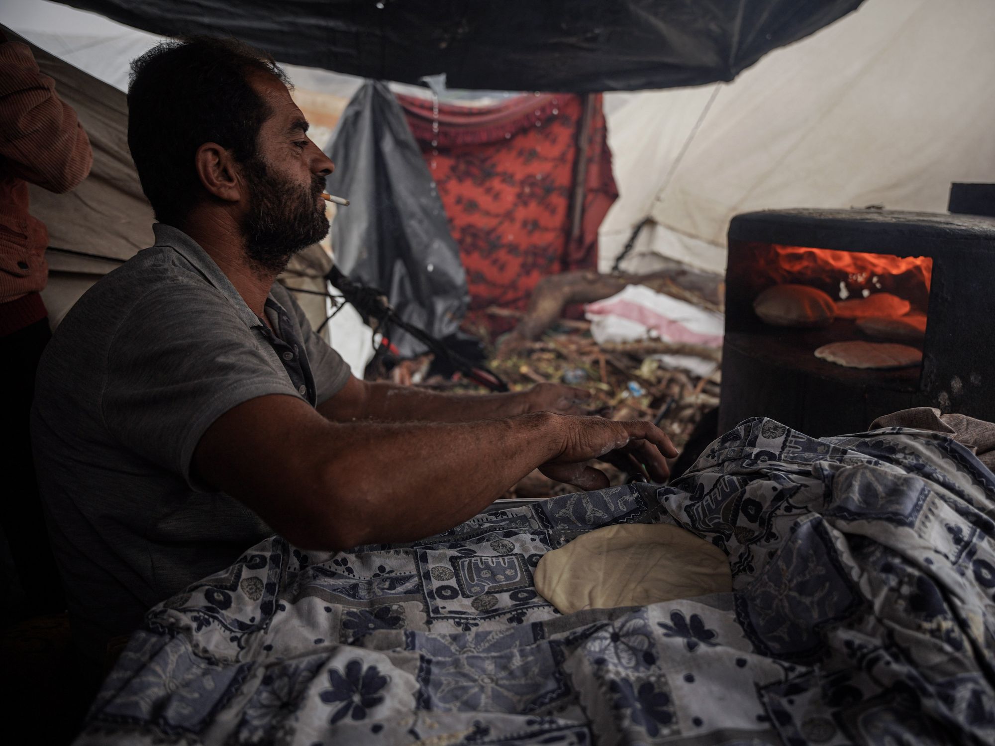 ​A Palestinian man bakes bread on a clay oven and firewood in a displacement camp in Khan Younis.