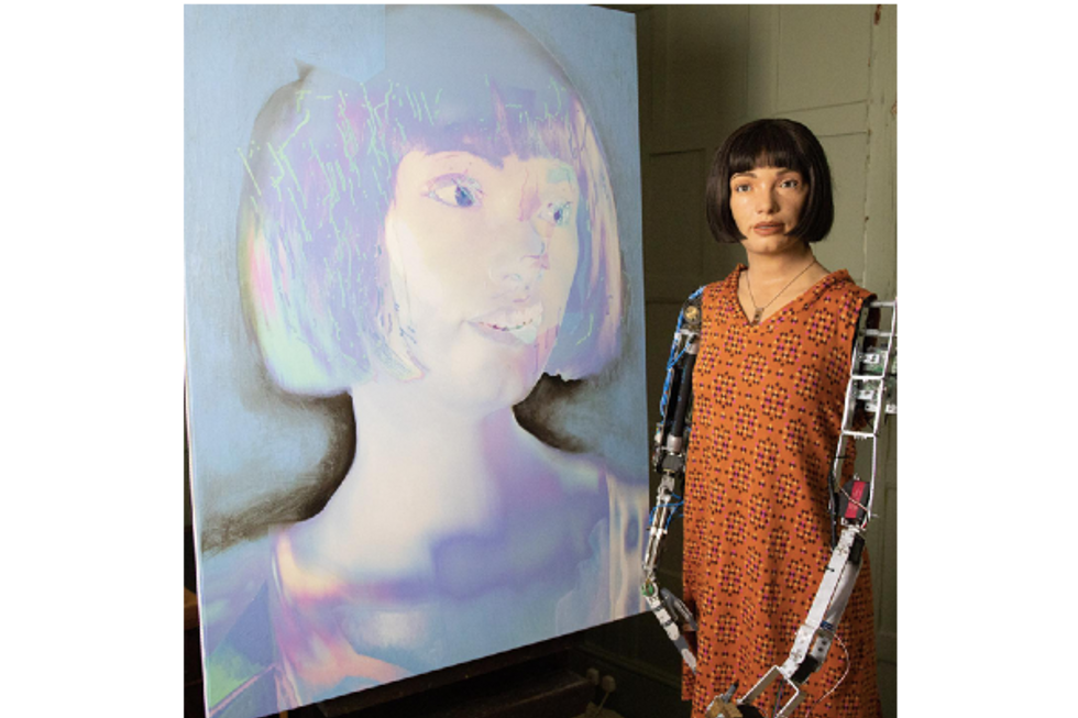 ​Photo of Ai-Da standing next to her self-portrait