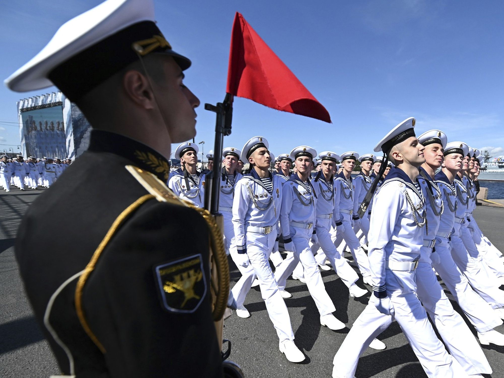 ​Russian naval sailors march past the review stand during Navy Day celebrations at Senate Square. Naval sailors are wearing white while a soldier is wearing a black uniform.
