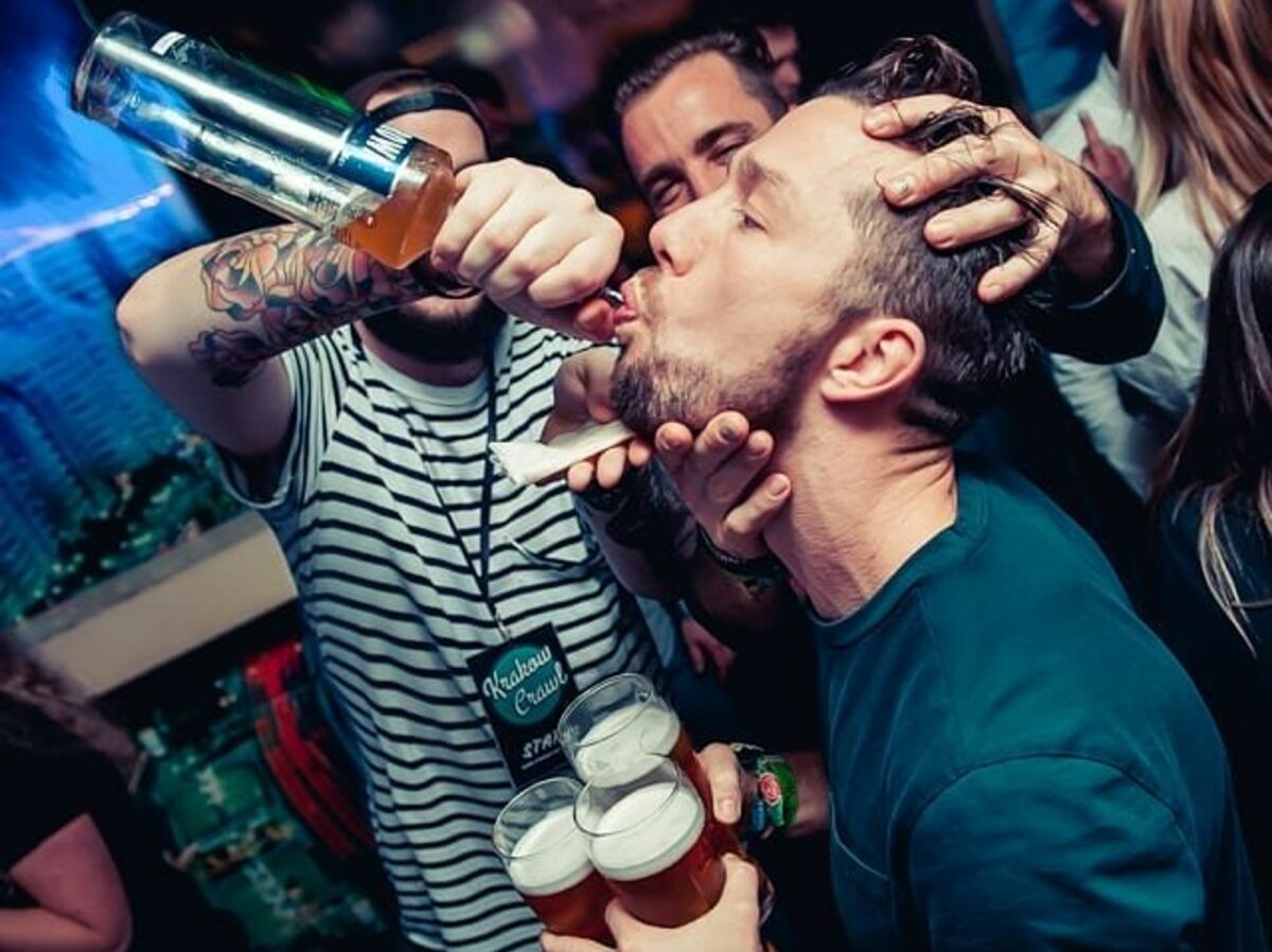 ​Tourist participating in a Krakow pub crawl. A man is holding three beers and drinking from a bottle his friend is pouring in his mouth.