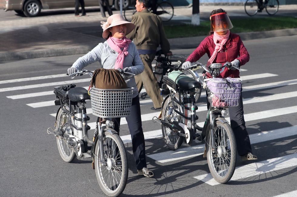 ​Two Women carry their bicycles with full baskets as they cross a street in the city of Pyongyang.
