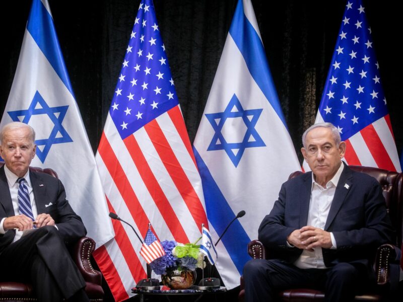 ​U.S President Biden looking on during a meeting with Israeli Prime Minister Netanyahu. In the background are a line of American and Israel flags.