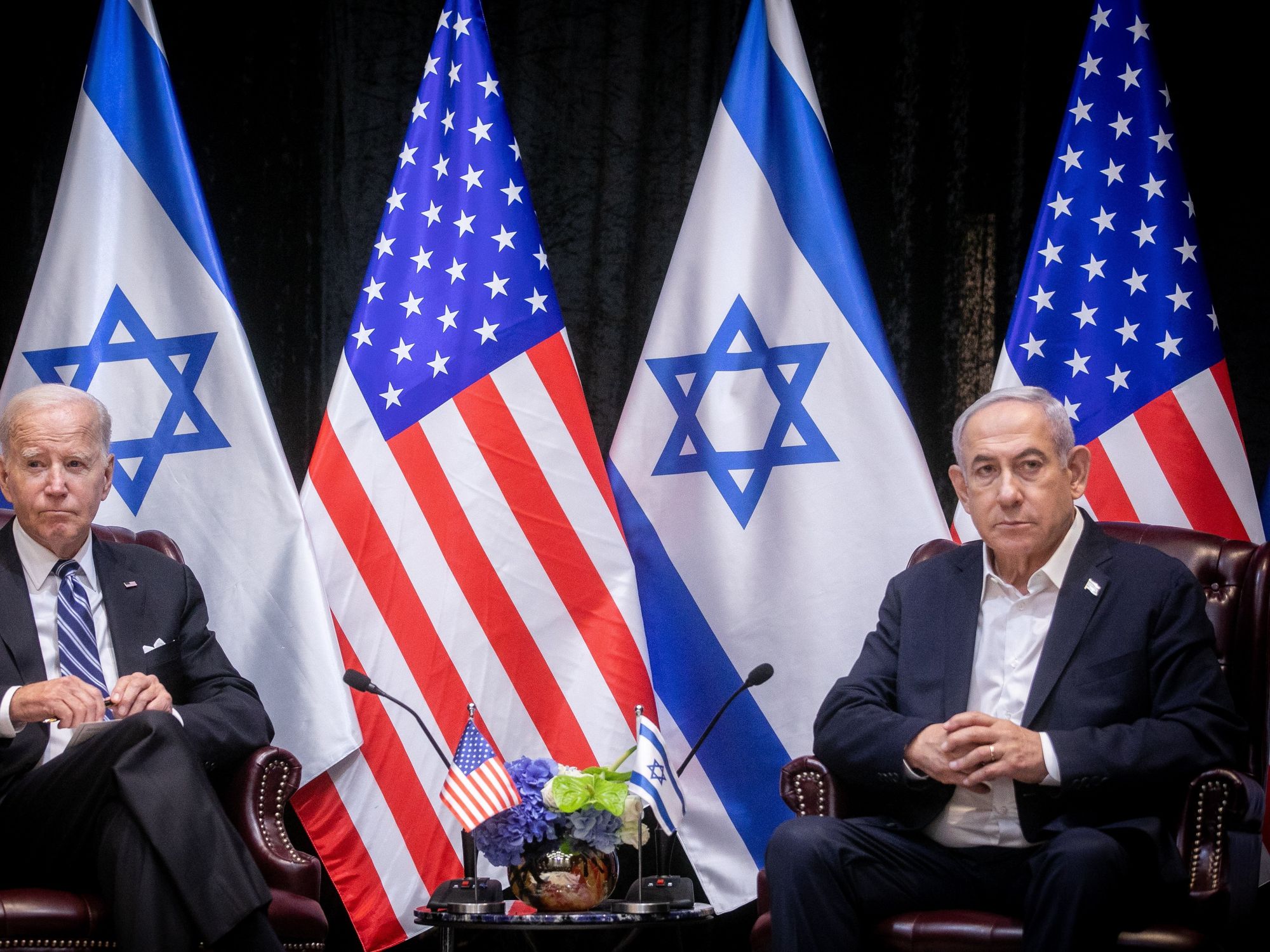 ​U.S President Biden looking on during a meeting with Israeli Prime Minister Netanyahu. In the background are a line of American and Israel flags.