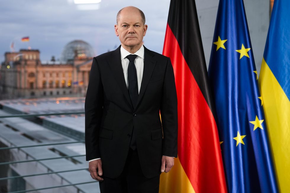German Chancellor Olaf Scholz standing in front of the German, EU and Ukrainian flags.