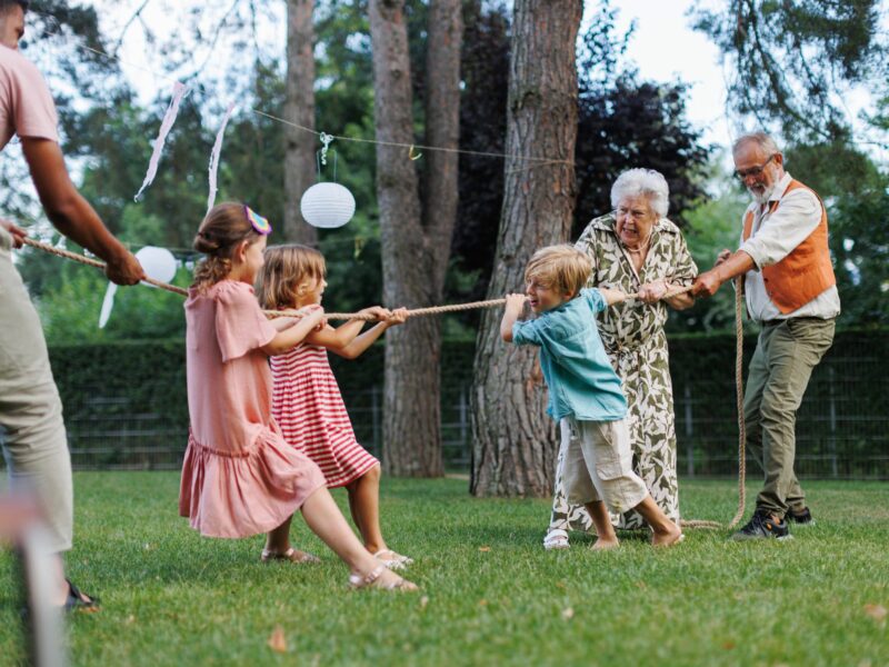 Grandparents and grandchildren play tug of war in a green yard with trees and baloons