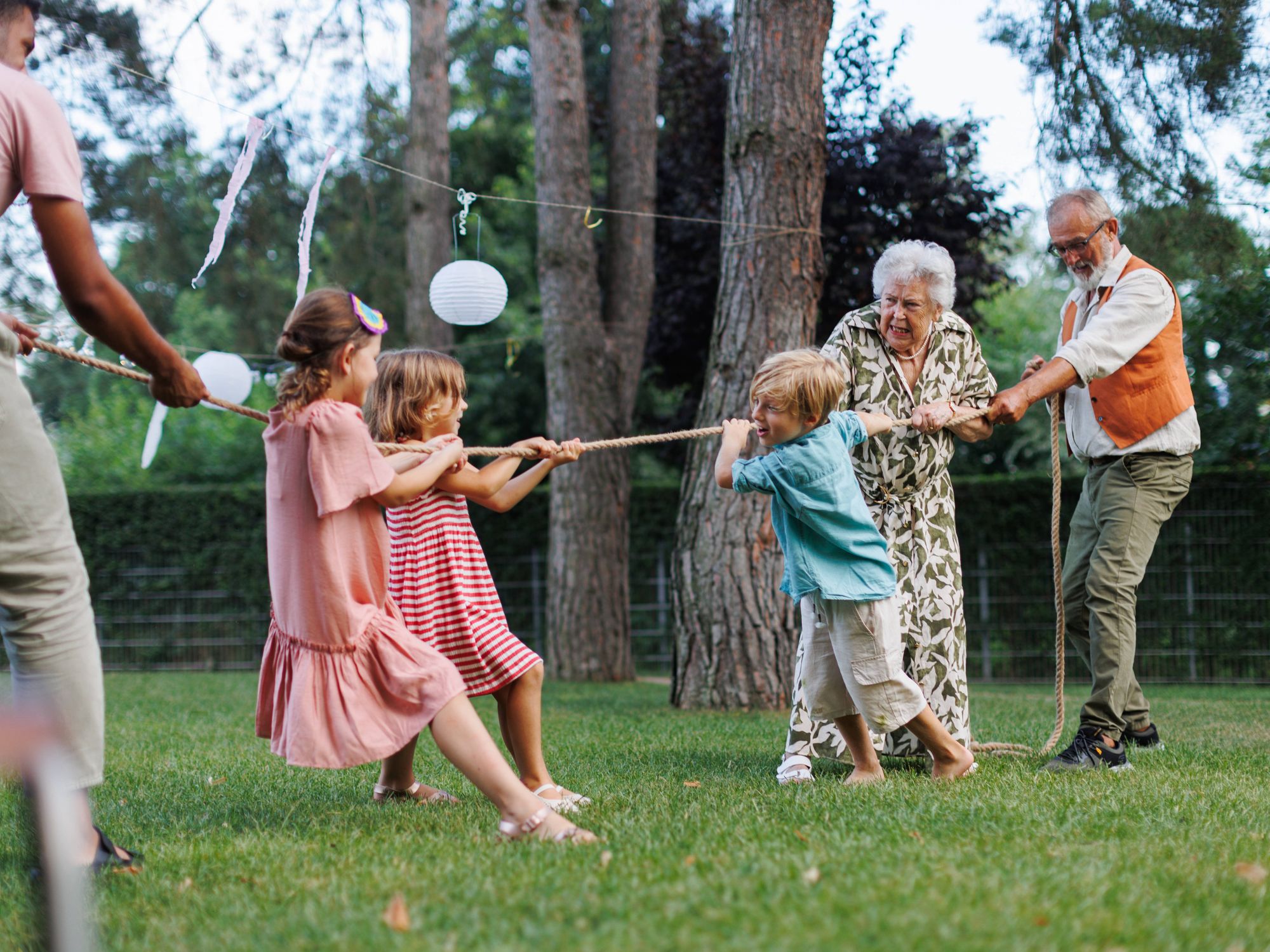 Grandparents and grandchildren play tug of war in a green yard with trees and baloons