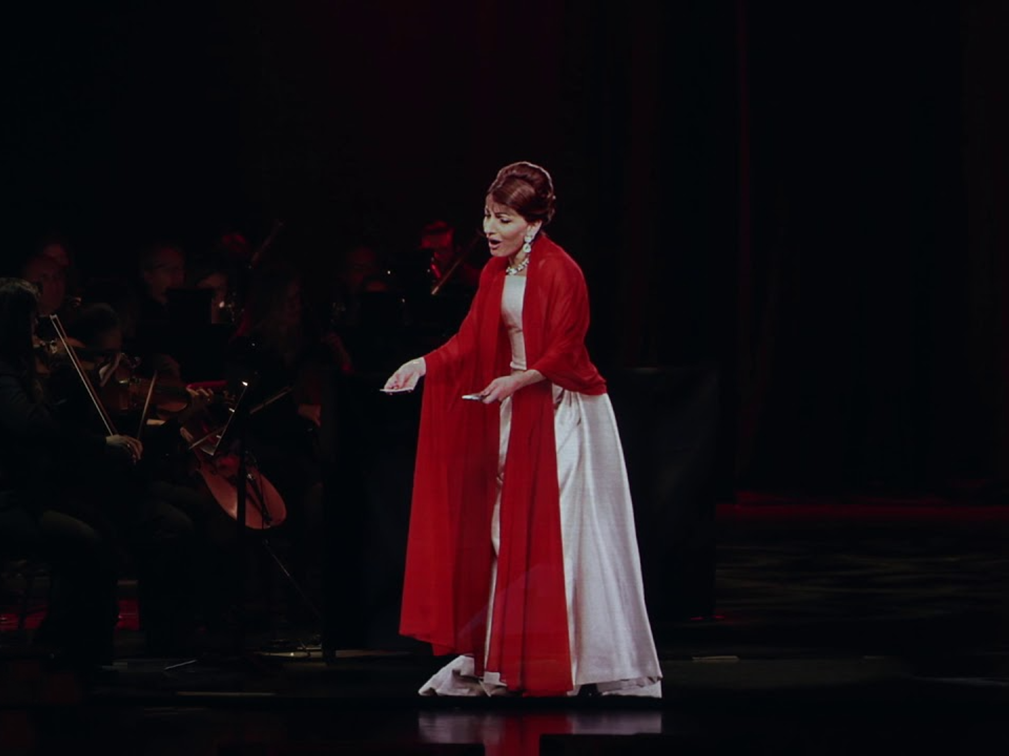 Hologram of Maria Callas wearing a white dress and red scarf peroming along side of an orchestra.