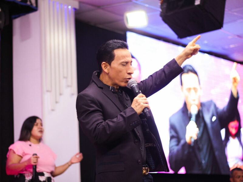 Image of a man in a suit, Esteban Acosta, a self-proclaimed apostle, giving a speech at ​La Unción Christian Community Church, a big screen behind him projecting his speech.