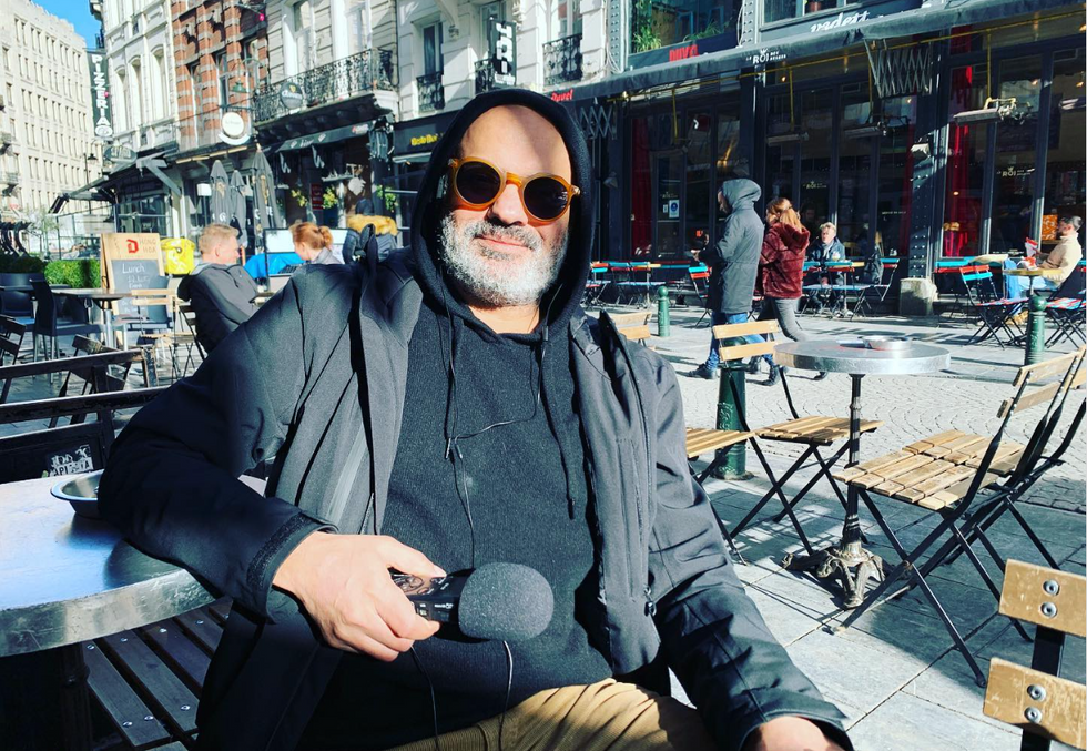 Image of a man wearing sunglasses sitting on the chair of a coffee shop outside and holding a microphone.