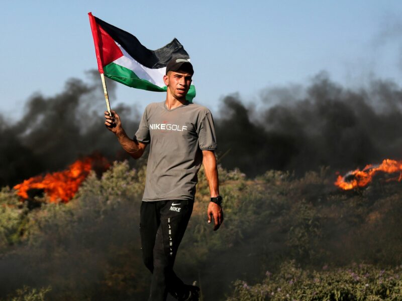Image of A Palestinian holding a national flag amidst burning tires near the border fence with Israel