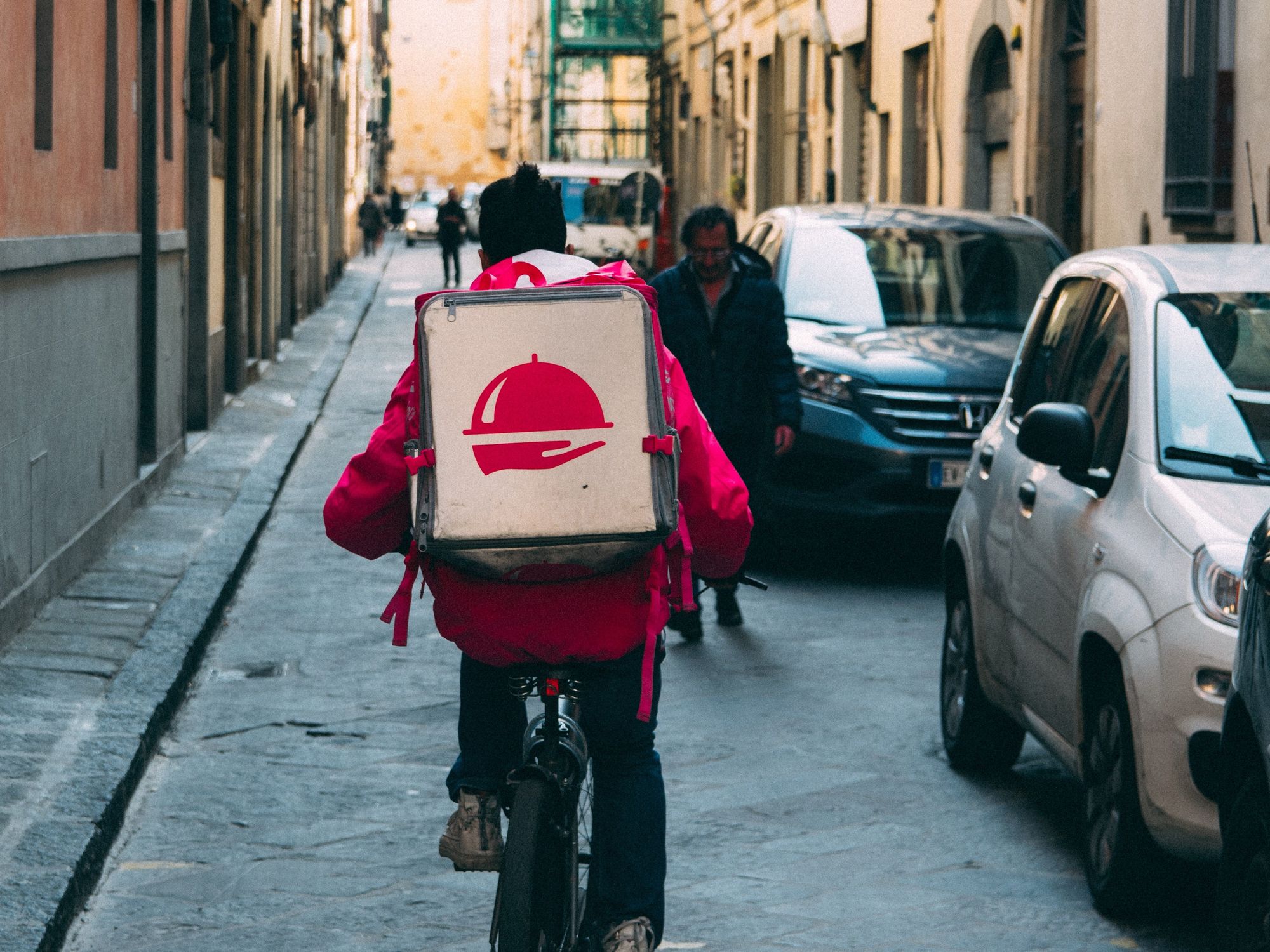 Image of a person riding a bike, on their way to deliver food in a street with cars and people.