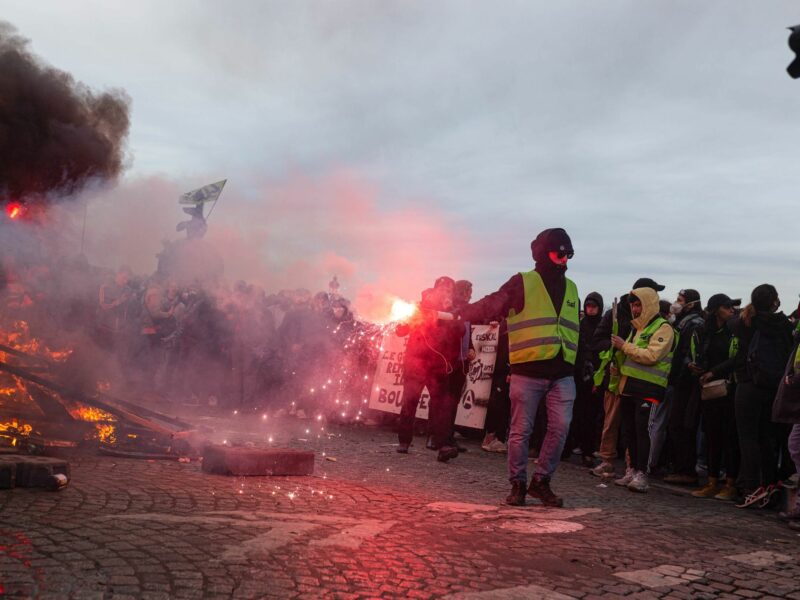 Image of a protester wearing a yellow vest and holding a smoke bomb near a fire started during a protest.
