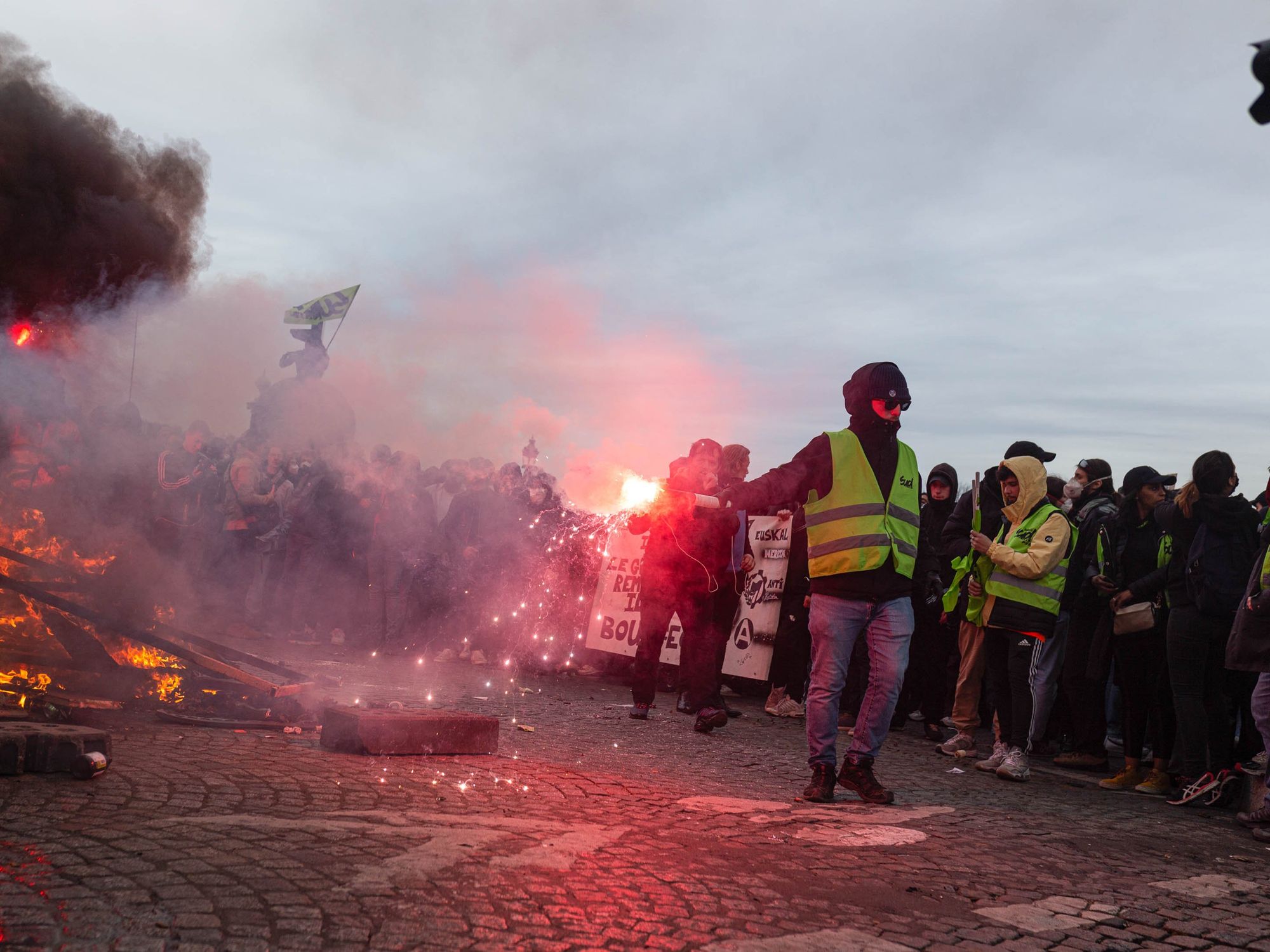 Image of a protester wearing a yellow vest and holding a smoke bomb near a fire started during a protest.
