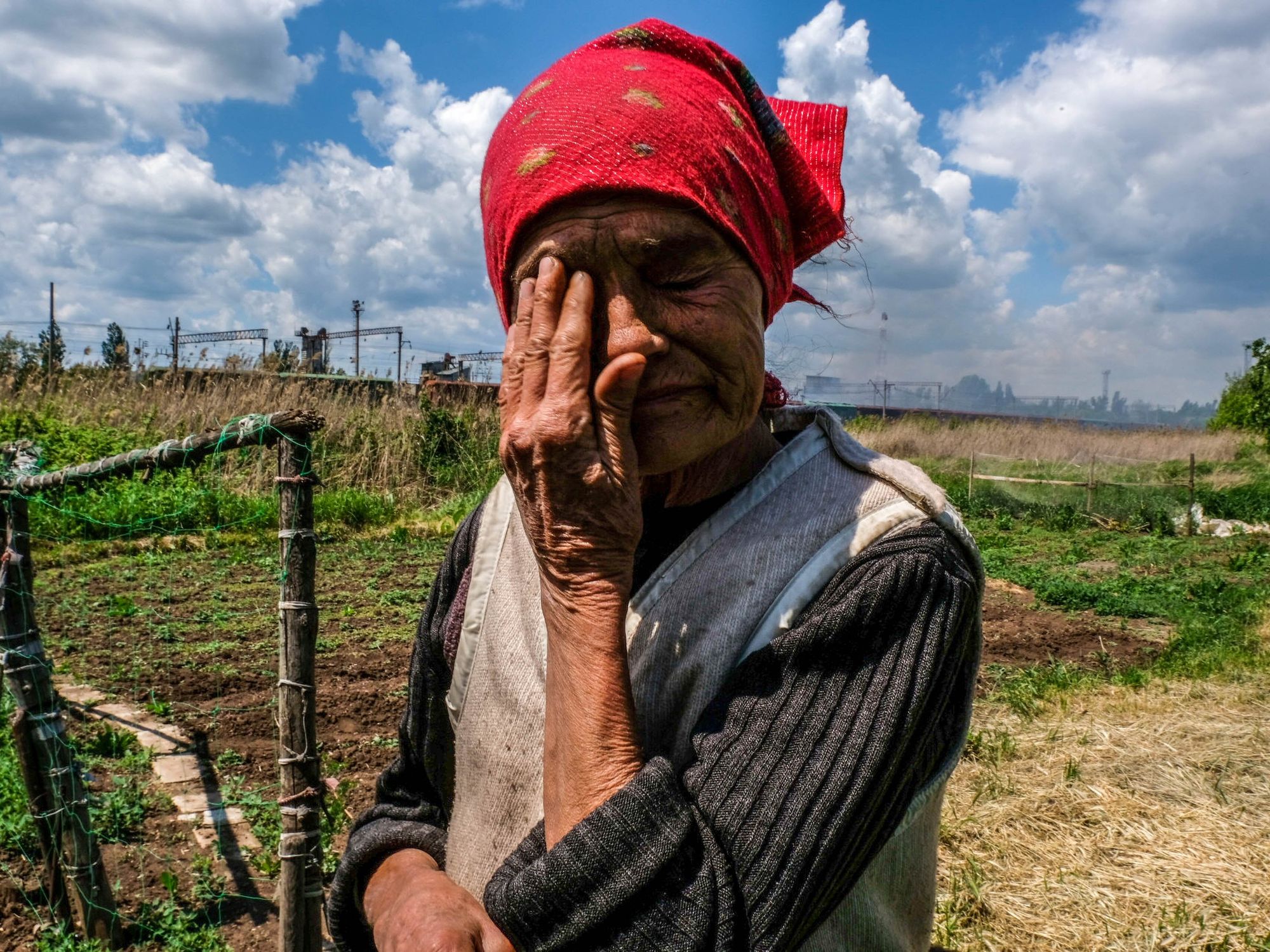 Image of an elderly woman in the countryside wiping hear tears with her hand, under a sky filled with smoke due to the bombing of a train station