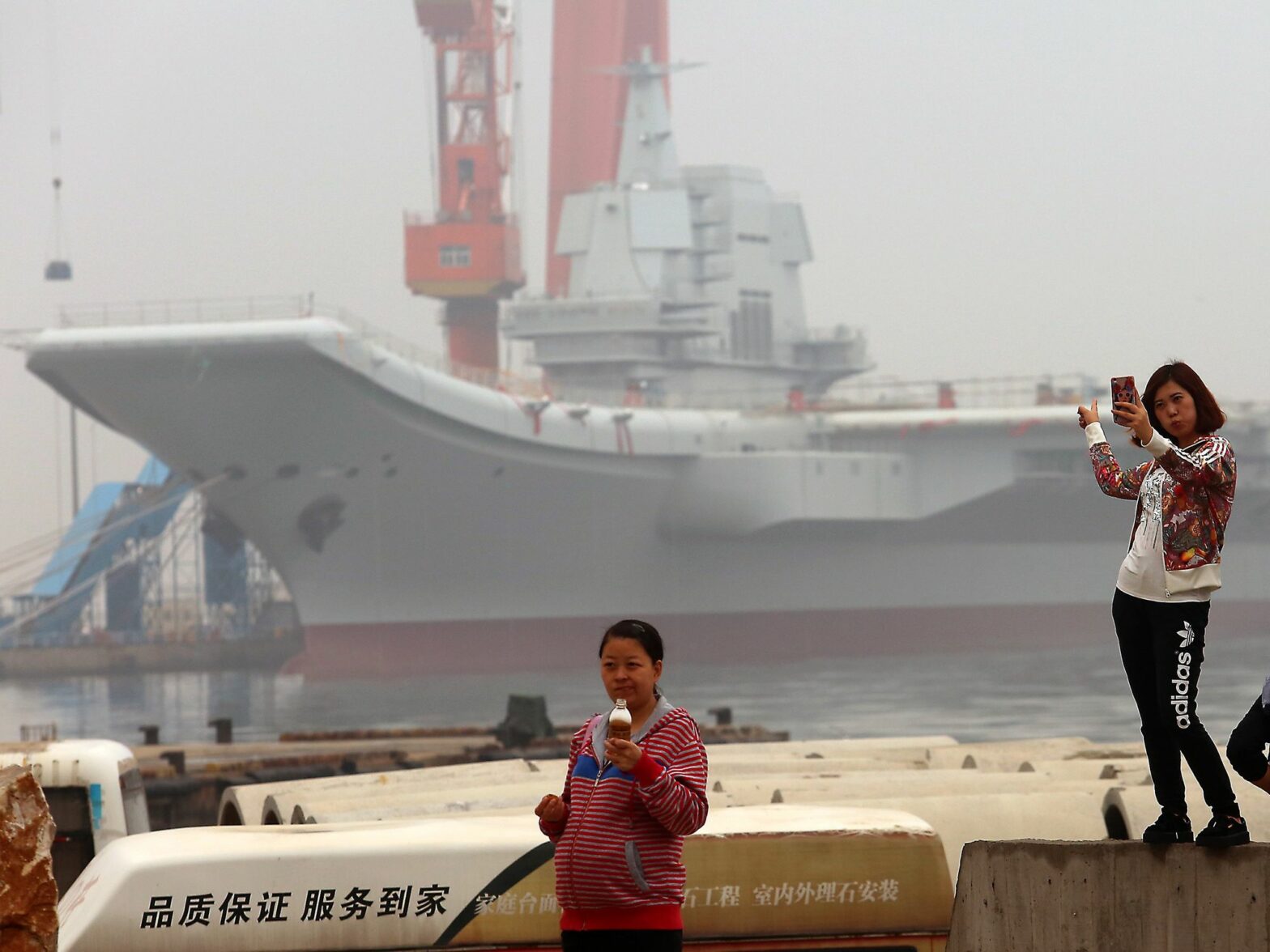 Image of Chinese girls taking selfies with China's second aircraft carrier, currently called the 001A, parked in a shipyard in the port city of Dalian in Liaoning Province, China.