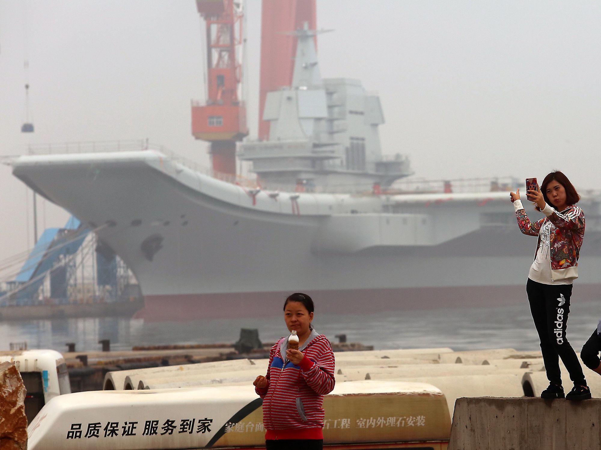 Image of Chinese girls taking selfies with China's second aircraft carrier, currently called the 001A, parked in a shipyard in the port city of Dalian in Liaoning Province, China.