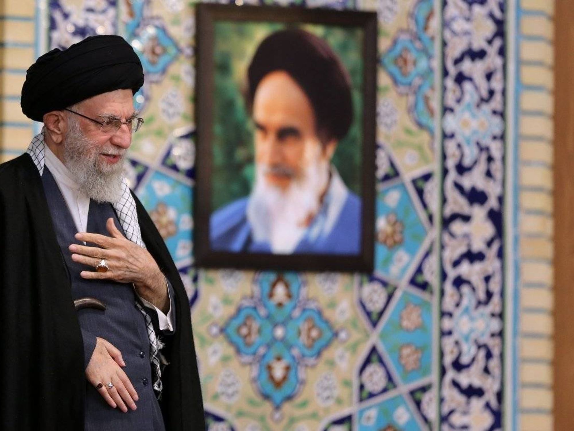 Image of Iran's supreme leader Ayatollah Khamenei smiling, a portrait of himself behind him.