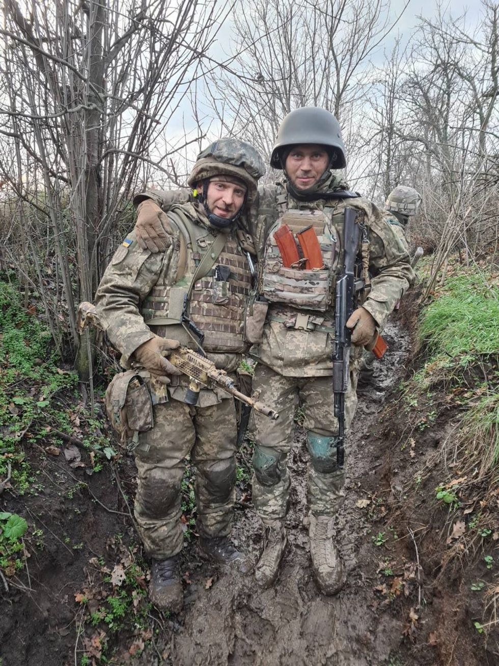 Image of two Ukrainian soldiers, armed and in uniform, standing next to each other, smiling at the camera.