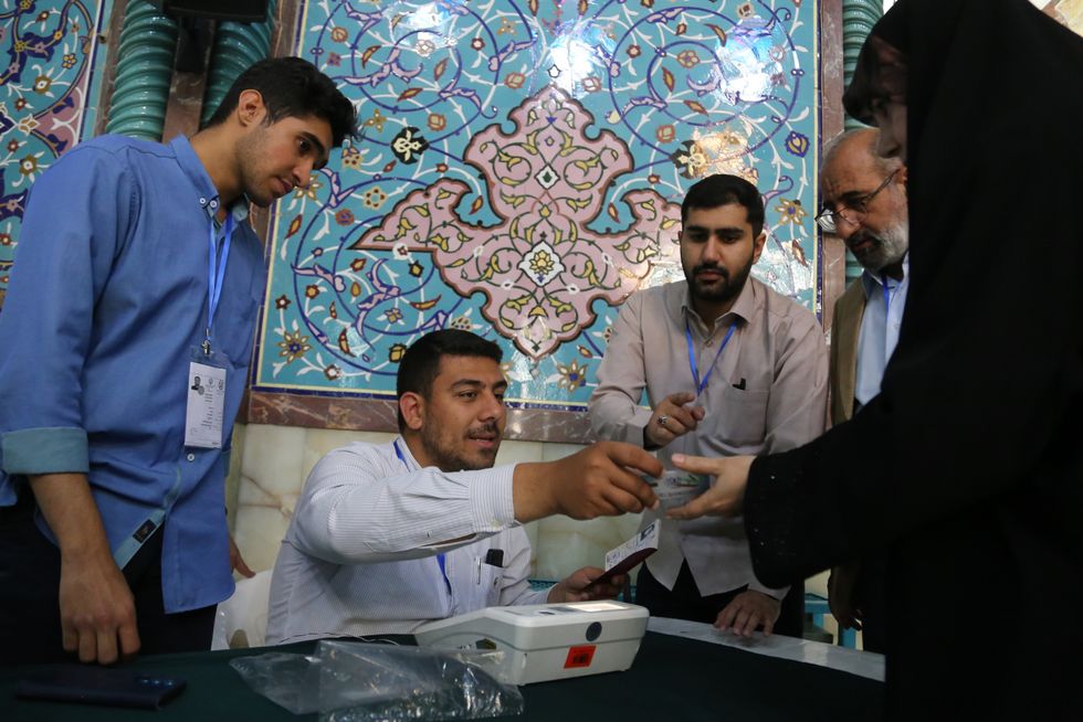 Iranian women in the process of voting and 4 men around the table with her as one of the men hand her something
