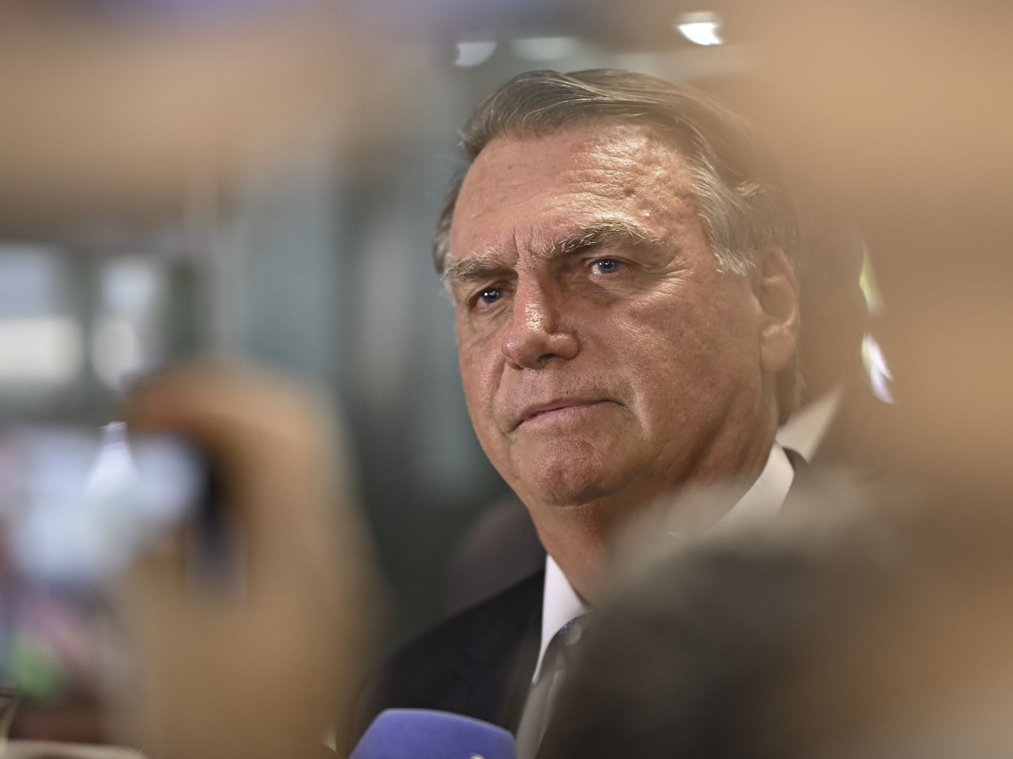 Jair Bolsonaro, in a suit, is seen through the hands and cameras of people taking his picture at a press conference