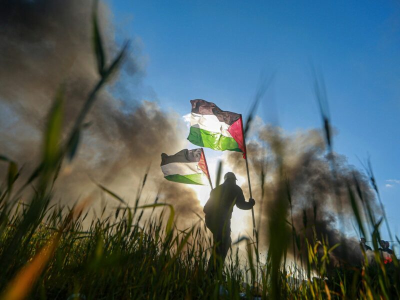 Low-angle shot of a man walking through a field carrying two Palestinian flags, with smoke in the background
