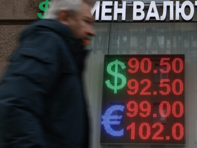 Man walking infront of a building with russian words on it and a sign displaying numbers in red for doallars as well as pounds