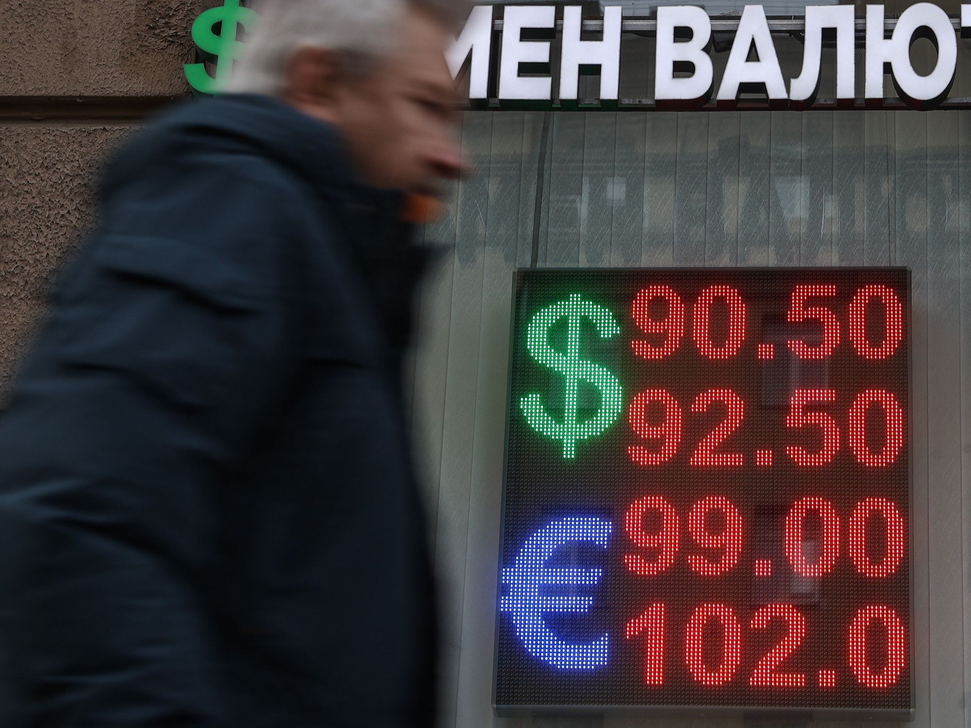 Man walking infront of a building with russian words on it and a sign displaying numbers in red for doallars as well as pounds
