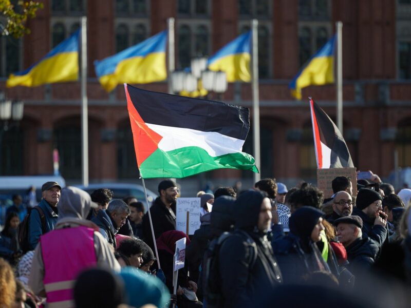 Many people stand outside holding Palestinian Flags with Ukrainian flags in the background