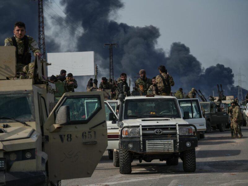 Men in camouflage ride in the backs of trucks with guns as pillars of black smoke stretch into the sky behind them.