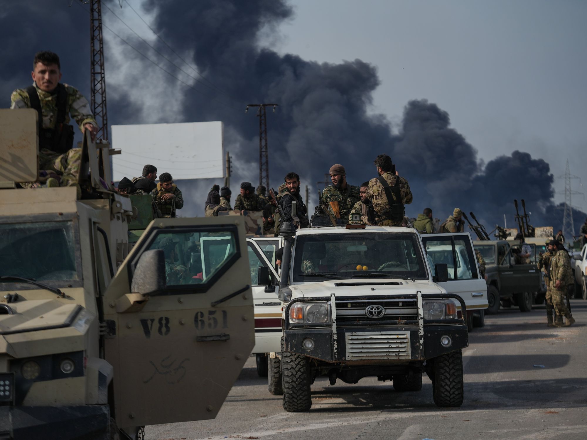 Men in camouflage ride in the backs of trucks with guns as pillars of black smoke stretch into the sky behind them.