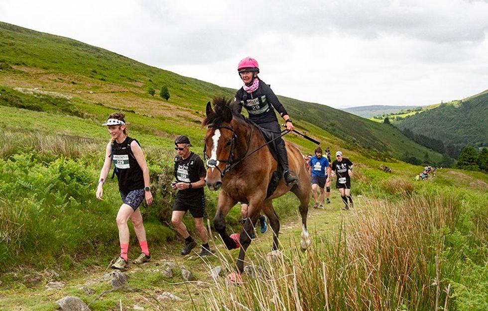 Participants of the man v.s. horse race​ caught in a photograph as the travers a hill. most are on foot bur one woman races in front of them with a horse.