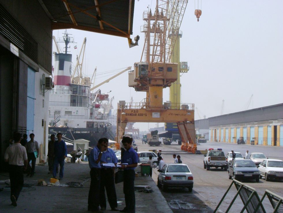 People and cars mill about in front of a crane in the port of Shahid Bahonar