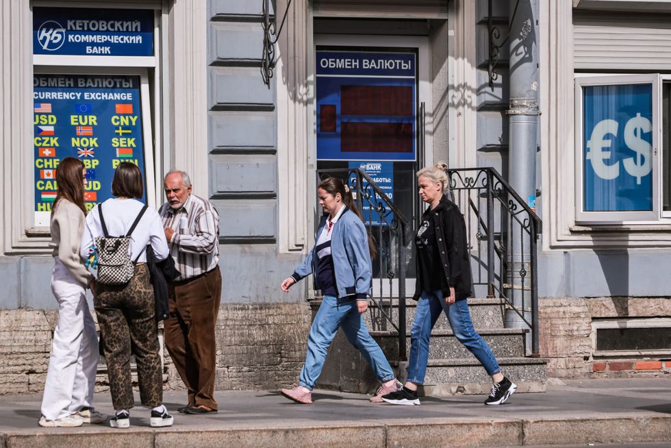 People infront of a buildiing with russian words above the door and window, and in one window a pound sign and the other some flags and above that in english "Currency Exchange