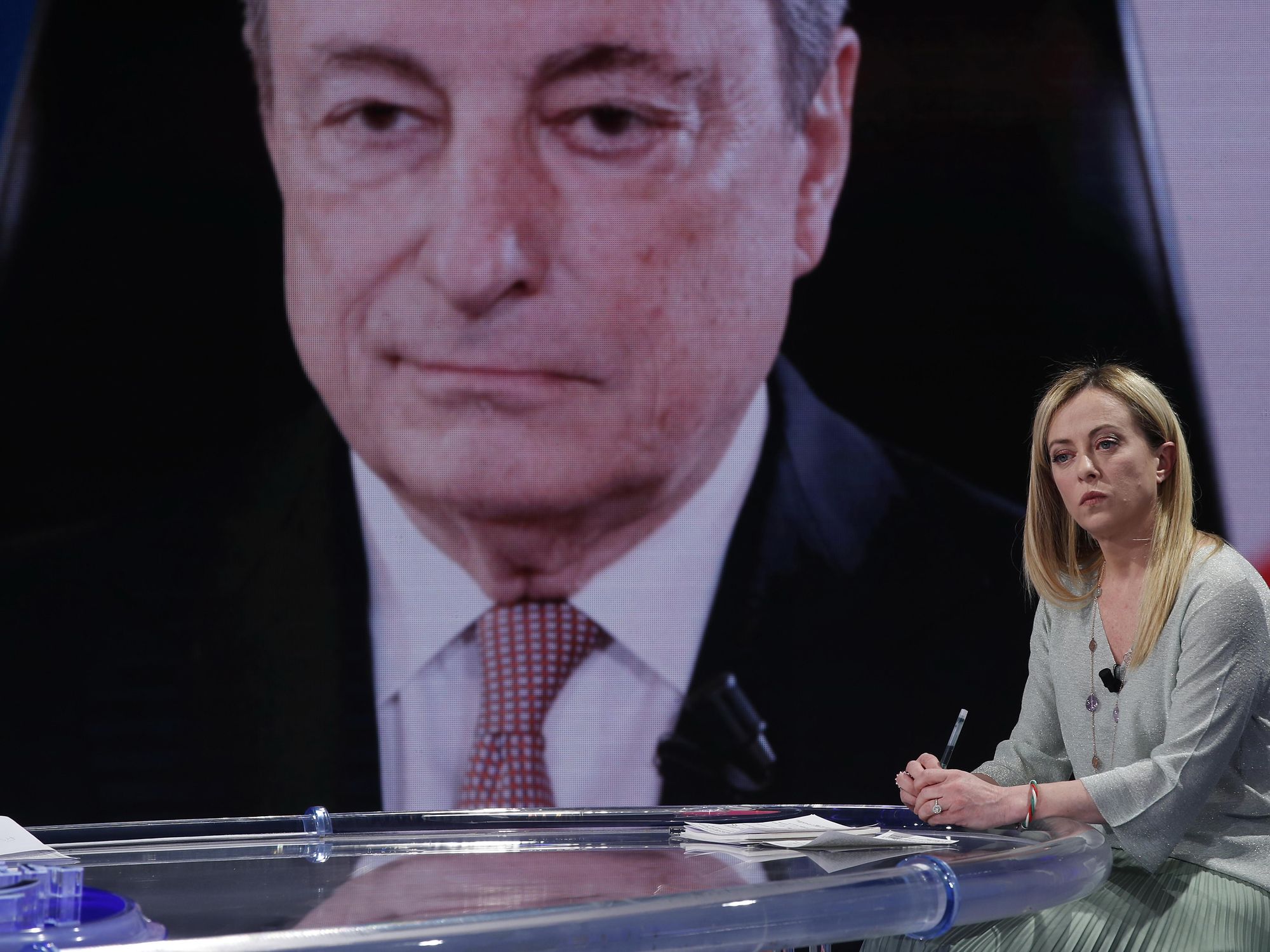 photo giorgia meloni on a tv set with mario draghi's image behind her