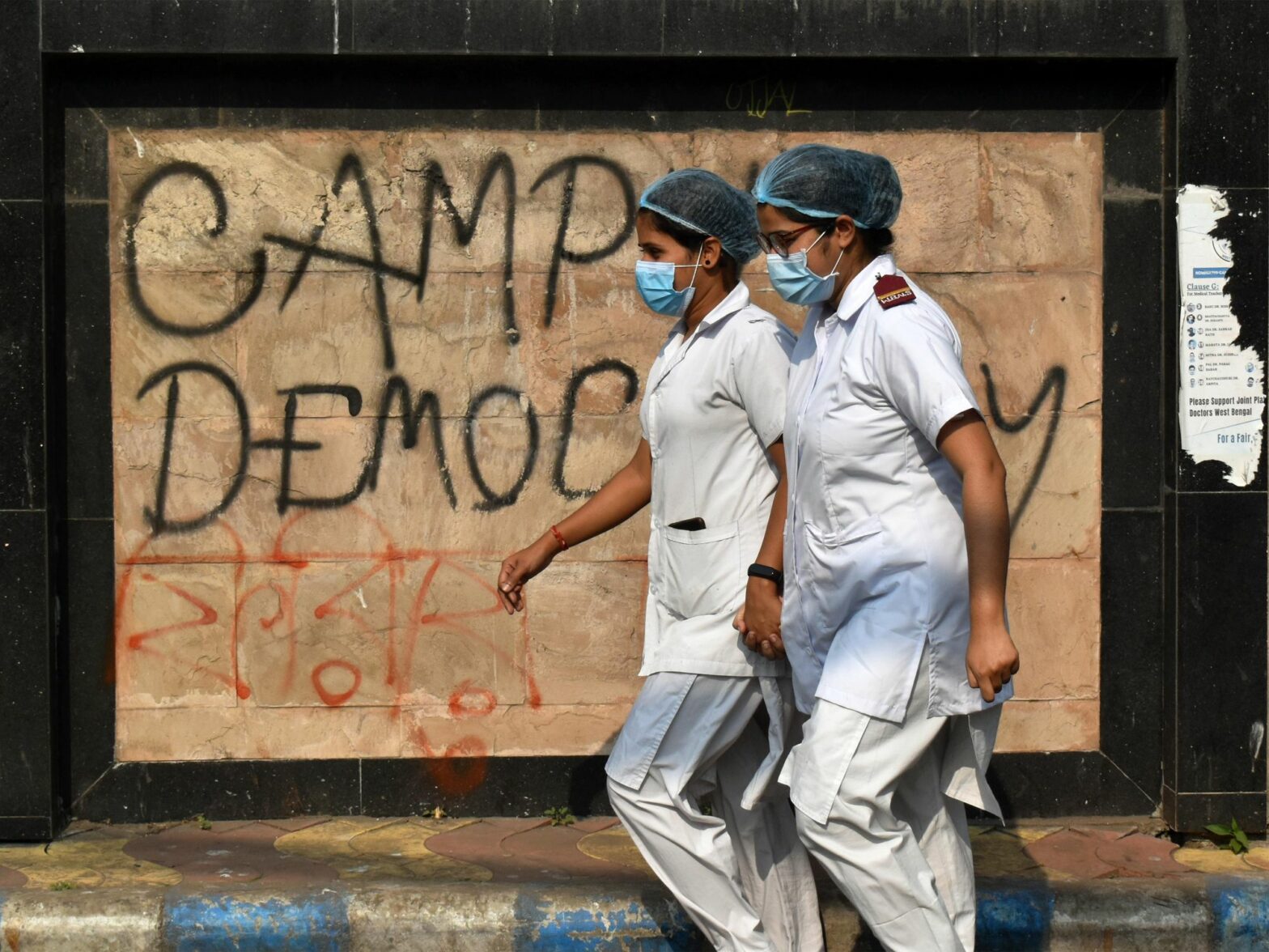 photo of 2 nurses in india walking past graffiti that says "democracy'