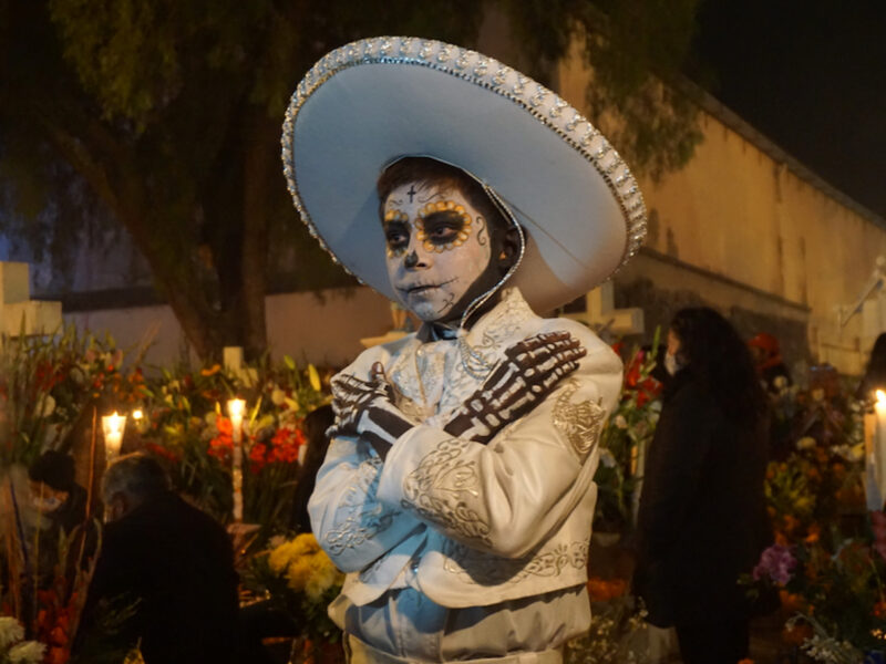 Photo of a boy black-and-white skeleton makeup, in white traditional Mexican clothes, during a Dia de Muertos celebration in Mexico City